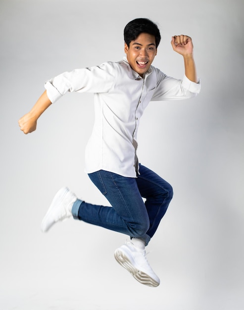 Retrato de um jovem asiático animado de camisa branca pulando enquanto celebra o sucesso no fundo cinza