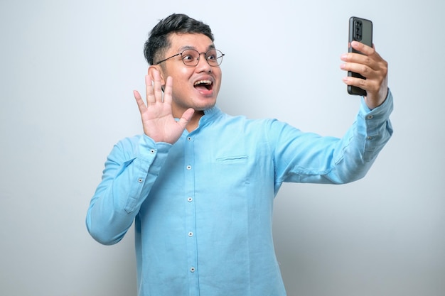 Retrato de um jovem asiático animado e alegre usando óculos e camisa casual usando telefone celular tirando selfie