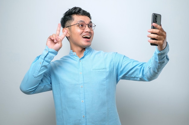 Retrato de um jovem asiático animado e alegre usando óculos e camisa casual usando telefone celular tirando selfie