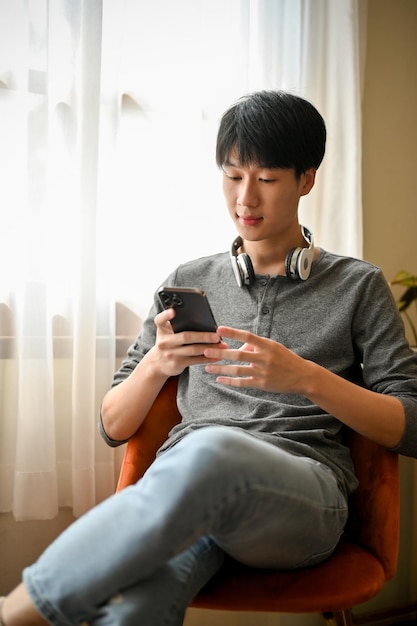 Retrato de um jovem asiático bonito usando seu smartphone em uma cadeira relaxando na sala de estar