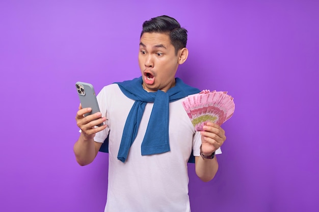Retrato de um jovem asiático chocado usando telefone celular e segurando notas de dinheiro isoladas sobre fundo roxo
