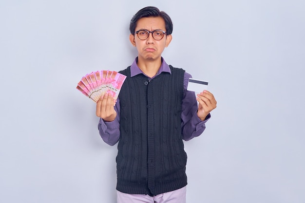 Retrato de um jovem asiático confuso em roupas de camisa segurando dinheiro em notas de rupia e mostrando cartão de crédito isolado no fundo branco Conceito de estilo de vida de pessoas