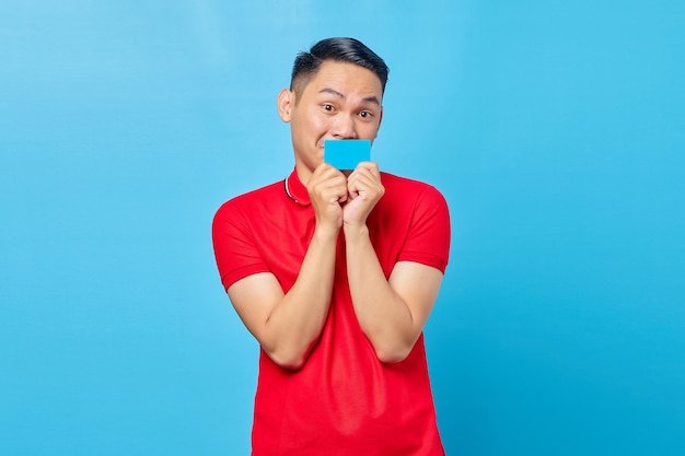 Retrato de um jovem asiático excitado e atraente cobrindo a boca com cartão de crédito e olhando para a câmera sobre fundo azul