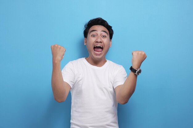 Retrato de um jovem asiático feliz e entusiasmado mostra um gesto de vitória celebrando a vitória sorrindo