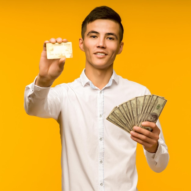 Retrato de um jovem asiático feliz segurando cartão de crédito e dinheiro na mão, sorrindo e olhando para a câmera em fundo amarelo isolado, sentindo-se positivo e aproveitando a imagem