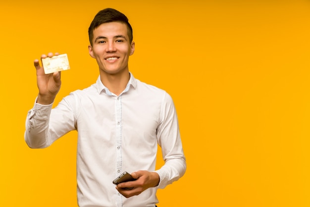 Foto retrato de um jovem asiático feliz segurando um cartão de crédito e falando ao telefone