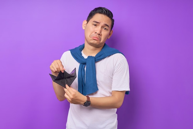 Retrato de um jovem asiático infeliz vestindo roupas casuais mostrando carteira vazia isolada em fundo roxo