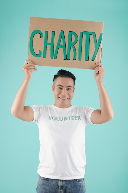 Retrato de um jovem asiático sorridente bonito segurando um cartaz com a inscrição de caridade e olhando para a câmera