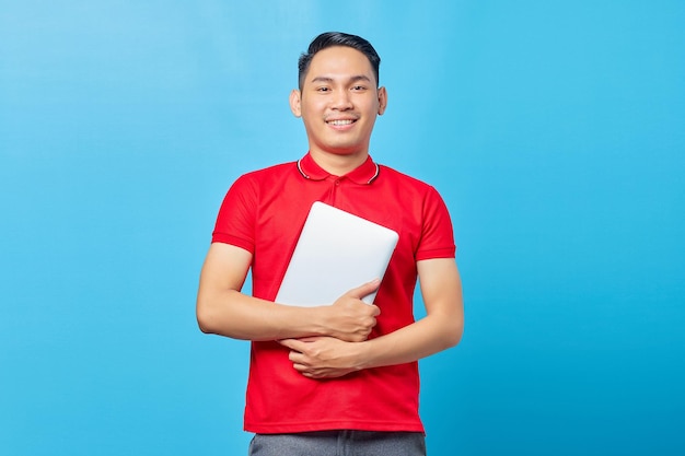 Retrato de um jovem asiático sorridente e bonito de camisa vermelha abraçando laptop isolado em fundo azul