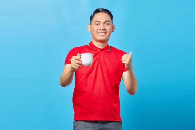 Retrato de um jovem asiático sorridente segurando uma xícara de café e apontando o dedo para o espaço de cópia isolado em fundo azul