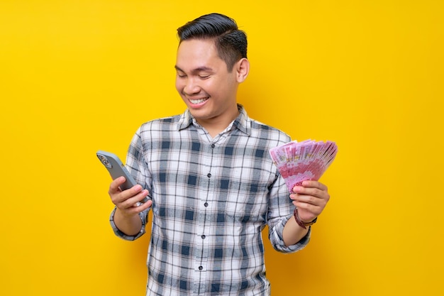 Retrato de um jovem asiático sorridente vestindo camisa xadrez segurando telefone celular e dinheiro em notas de rupia isoladas no conceito de estilo de vida de pessoas de fundo amarelo