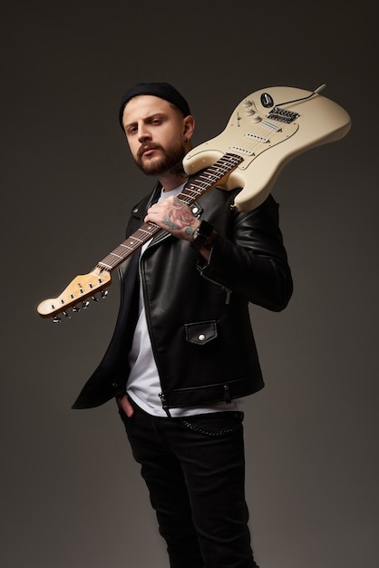 Retrato de um jovem atraente em uma jaqueta de couro e tatuagens com uma guitarra elétrica no ombro. fundo escuro