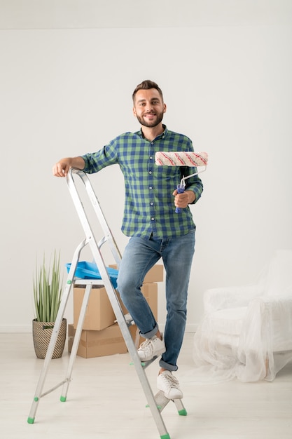 Retrato de um jovem barbudo alegre parado com rolo de pintura na escada em um apartamento novo com coisas em movimento