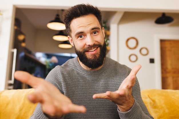 Foto retrato de um jovem barbudo biracial sorridente gesticulando enquanto está sentado no sofá da sala de estar. feliz, bonito, inalterado, estilo de vida, relaxamento e conceito de casa.