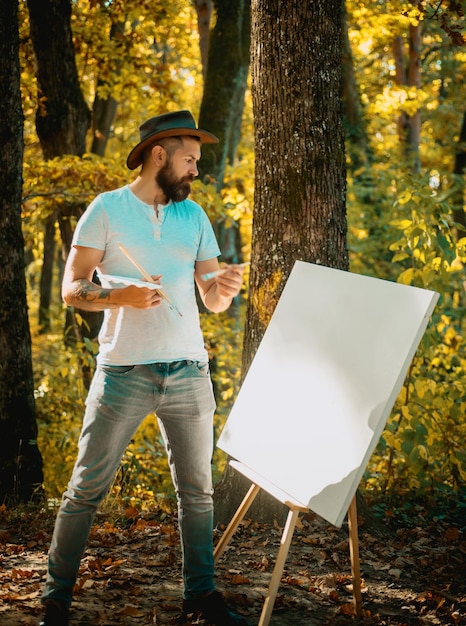 Foto retrato de um jovem barbudo de chapéu pintando um quadro com pincéis e cavalete na natureza do outono em um