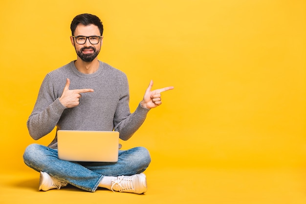 Retrato de um jovem barbudo feliz casual segurando o computador portátil enquanto está sentado no chão isolado sobre fundo amarelo. Dedo indicador.