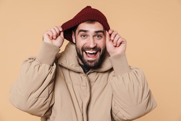 Retrato de um jovem barbudo feliz, vestindo uma jaqueta de inverno e chapéu em pé, isolado em bege