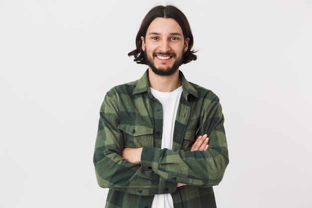 Retrato de um jovem barbudo homem moreno vestindo uma camisa xadrez em pé, isolado na parede branca, com os braços cruzados