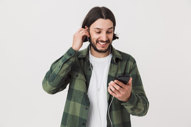 Retrato de um jovem barbudo homem moreno, vestindo uma camisa xadrez em pé, isolado na parede branca, ouvindo música com fones de ouvido, segurando um telefone celular