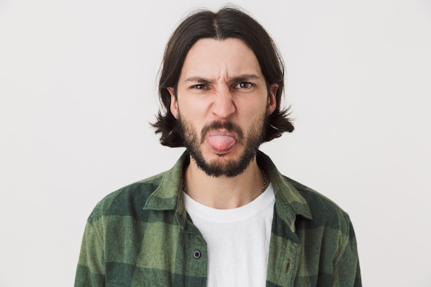 Retrato de um jovem barbudo moreno enojado, vestindo uma camisa xadrez, isolado sobre uma parede branca