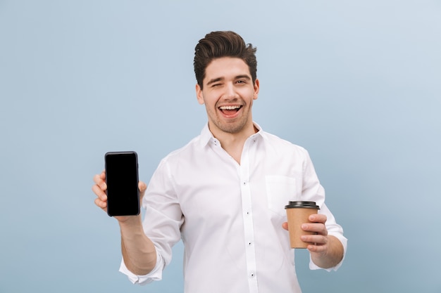 Retrato de um jovem bonito e alegre, isolado em um azul, segurando uma xícara de café para viagem, mostrando a tela em branco do celular