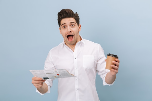 Retrato de um jovem bonito e alegre parado isolado em um azul, segurando uma xícara de café para viagem, lendo jornal