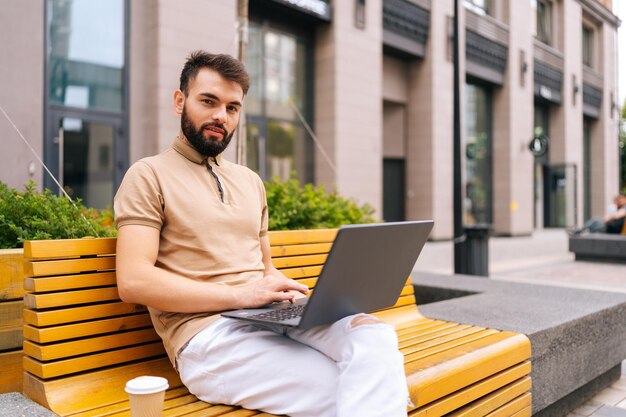 Retrato de um jovem bonito e barbudo trabalhando remotamente no computador portátil sentado no banco na rua da cidade e confiante olhando para a câmera