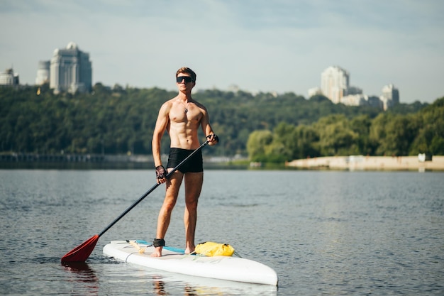 Retrato de um jovem bonito em óculos de sol e um torso musculoso em pé sobre uma placa de sup