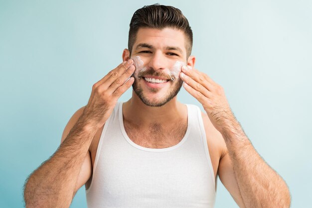 Retrato de um jovem bonito feliz aplicando hidratante no rosto em pé contra o fundo turquesa