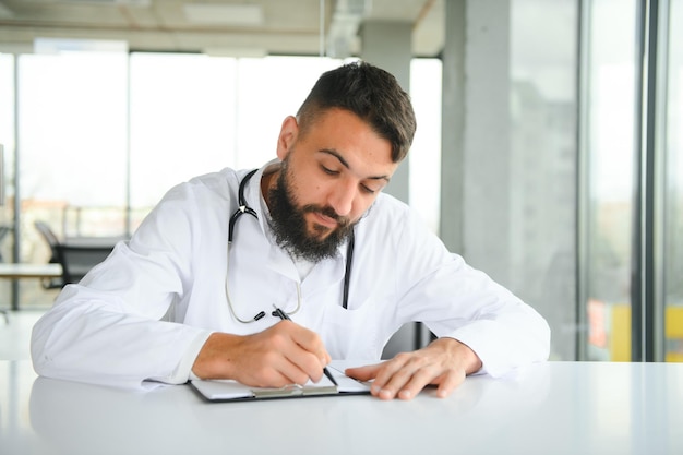 Retrato de um jovem bonito médico árabe em roupas de trabalho posando na clínica moderna sentado na mesa de trabalho