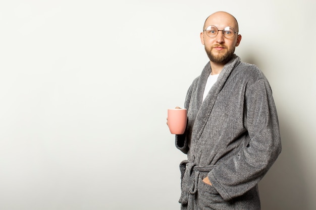 Retrato de um jovem careca com uma barba em um roupão e óculos, segurando uma xícara de café quente