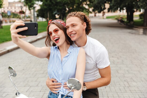 Retrato de um jovem casal alegre andando juntos em uma moto na rua da cidade, tirando uma selfie