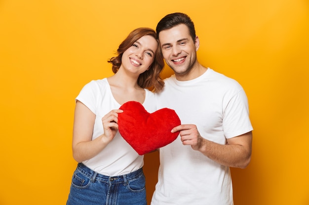 Retrato de um jovem casal alegre em pé, isolado na parede amarela, mostrando um coração vermelho