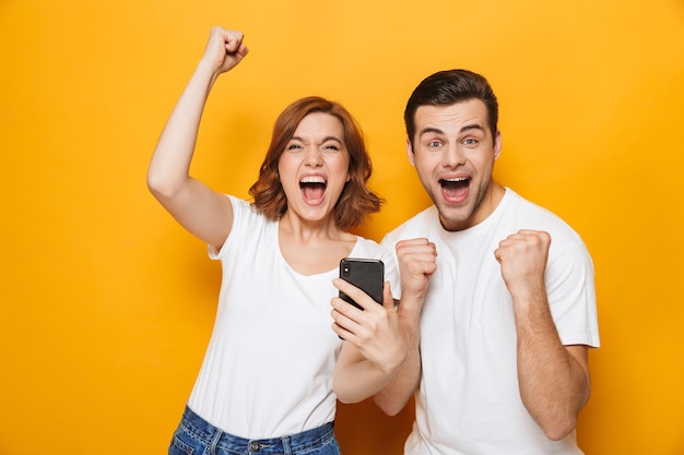 Retrato de um jovem casal alegre, isolado na parede amarela, usando telefones celulares, comemorando o sucesso