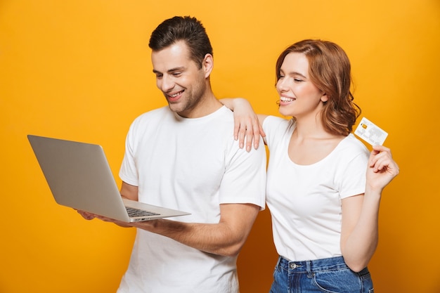 Retrato de um jovem casal alegre, isolado na parede amarela, usando um laptop para fazer compras online