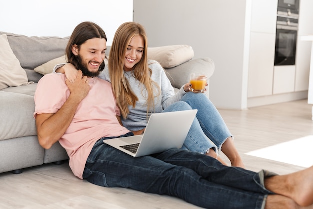 Retrato de um jovem casal apaixonado abraçando otimista perto do sofá, usando o computador laptop dentro de casa em casa.