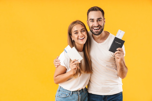 Retrato de um jovem casal atraente e alegre, vestindo roupas casuais, em pé, isolado na parede amarela, mostrando o passaporte com as passagens aéreas