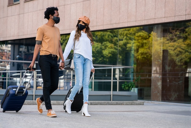 Foto retrato de um jovem casal de turistas usando máscara protetora e carregando mala enquanto caminha ao ar livre na rua. conceito de turismo. novo conceito de estilo de vida normal.
