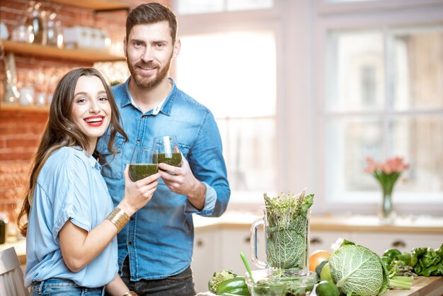 Retrato de um jovem casal de vegetarianos bebendo smoothie fresco na cozinha com comida verde saudável em casa