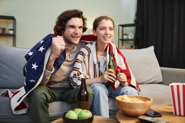 Retrato de um jovem casal emocional com a bandeira americana assistindo a uma partida esportiva em casa