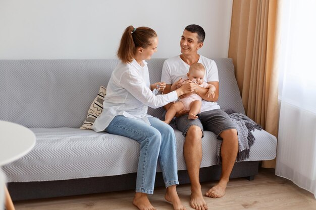 Retrato de um jovem casal encantado, vestindo roupas de estilo casual, sentado no sofá, na leve sala de estar com um bebê infantil, aproveitando para passar juntos, feliz fim de semana.