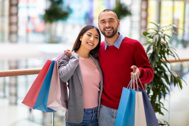 Foto retrato de um jovem casal feliz e diversificado posando com sacolas de compras sorrindo para a câmera no enorme shopping da cidade