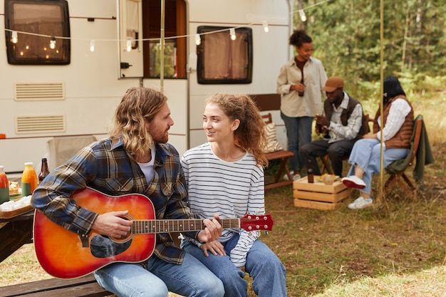 Retrato de um jovem casal feliz tocando violão enquanto desfruta de acampamento ao ar livre com a cópia da van de reboque.