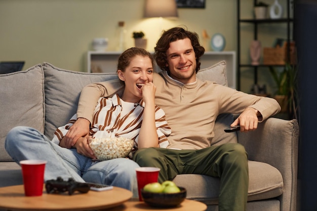 Retrato de um jovem casal feliz vendo filmes em casa e comendo pipocas sentados juntos no sofá