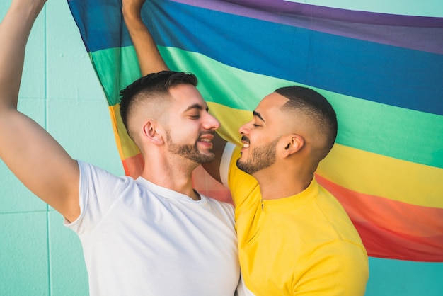 Foto retrato de um jovem casal gay, abraçando e mostrando seu amor com a bandeira do arco-íris na rua. lgbt e conceito de amor.