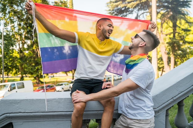 Retrato de um jovem casal gay abraçando e mostrando seu amor com a bandeira do arco-íris no stret. LGBT e conceito de amor.
