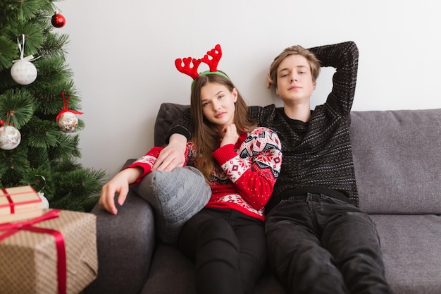 Retrato de um jovem casal lindo sentado no sofá em casa e sonhando olhando para a câmera com a árvore de Natal perto