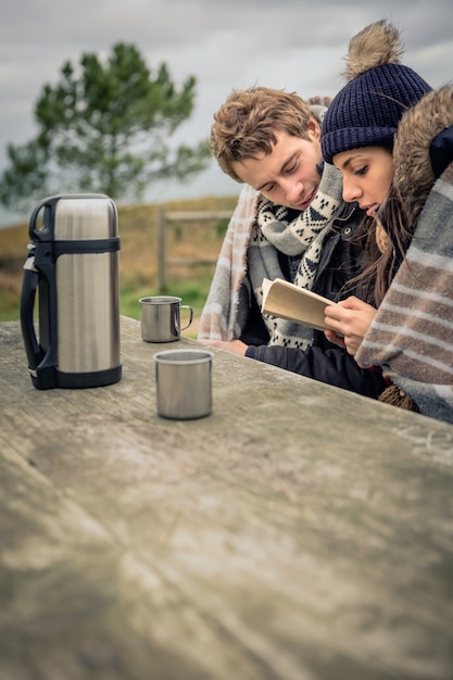 Retrato de um jovem casal lindo sob o cobertor lendo um livro em um dia frio com mar e céu escuro e nublado ao fundo.