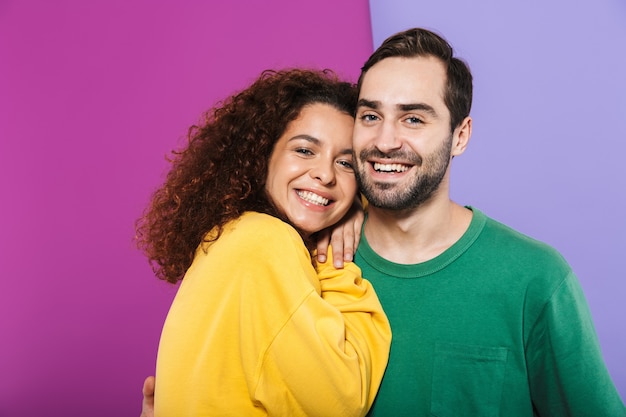 Foto retrato de um jovem casal moreno, um homem e uma mulher com roupas coloridas, sorrindo e se abraçando, isolados.