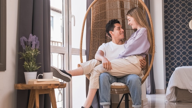 Retrato de um jovem casal olhando um para o outro enquanto estão sentados em uma cadeira de vime juntos em casa